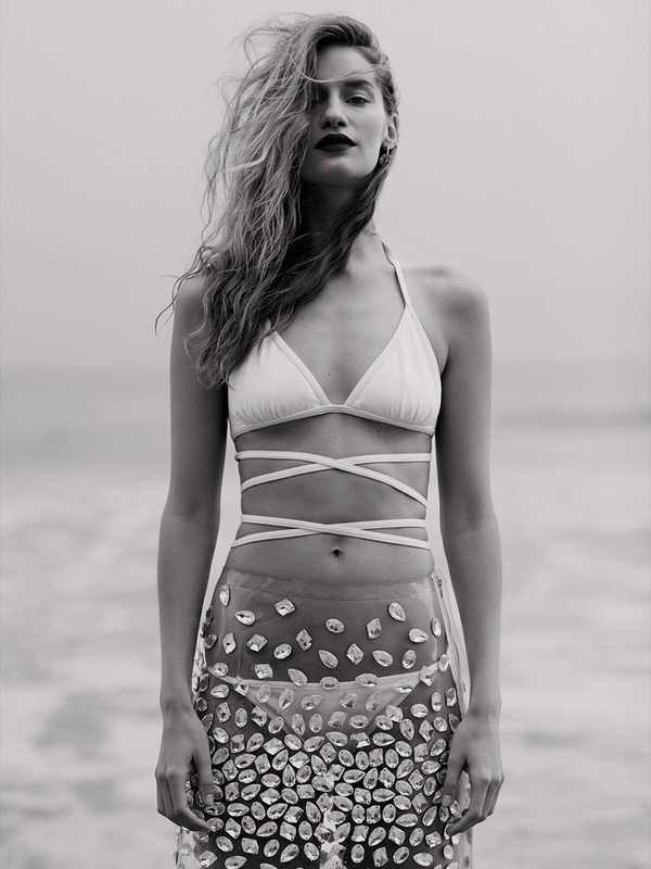 Monochrome portrait of a woman in a bikini top and embellished sheer skirt standing on a beach with soft lighting and ocean background.