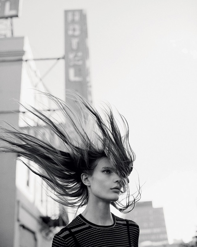 Monochrome outdoor fashion shot of a woman with windblown hair in front of a Hotel sign, wearing a striped top and gazing slightly down.