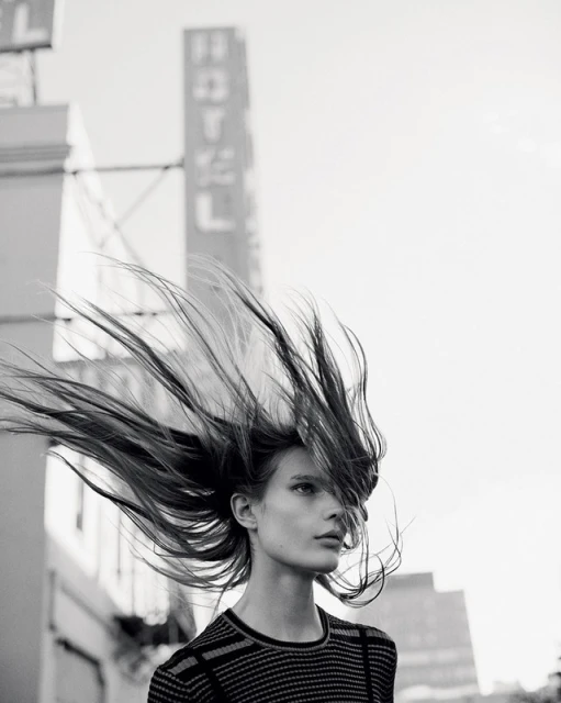 Monochrome outdoor fashion shot of a woman with windblown hair in front of a Hotel sign, wearing a striped top and gazing slightly down.