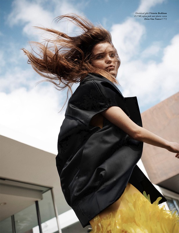 Fashion shot of an adult woman wearing a black jacket and yellow skirt, hair blowing in the wind, in front of a modern building and blue sky.