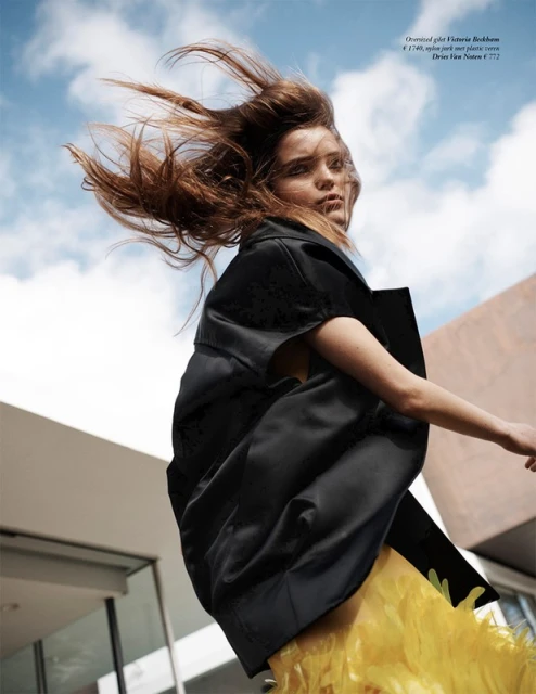 Fashion shot of an adult woman wearing a black jacket and yellow skirt, hair blowing in the wind, in front of a modern building and blue sky.
