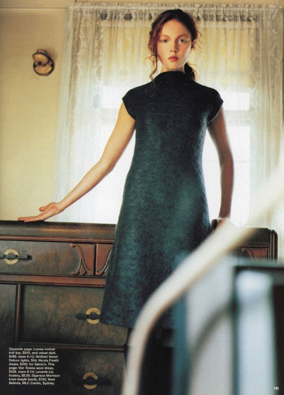Woman in a dark green vintage dress, indoors. She is standing next to a wooden dresser with brass pulls, with a window behind her.