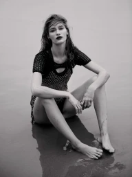 Monochrome portrait of a woman sitting barefoot on a wet sandy beach wearing a dark mesh dress styled by David Cox.