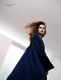 Model with long blowing hair wearing a deep blue pleated dress captured from a low angle against a white studio ceiling.