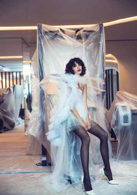 A woman in a white dress with a ruffled collar sits in an indoor space amid furniture covered in plastic sheeting. She wears sheer black stockings and white heels.
