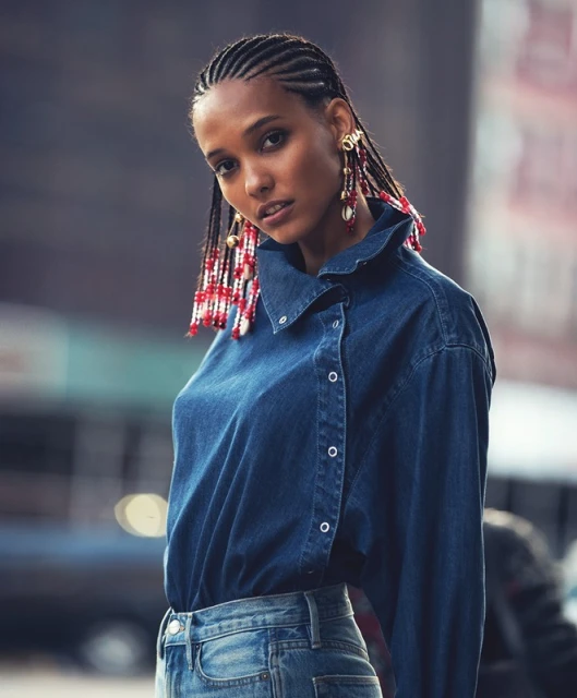Attractive Black woman in fashionable denim shirt and jeans with beaded cornrows poses outdoors against blurred urban background.