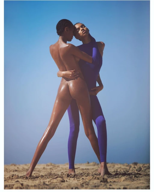 Two African American women embrace on a sandy beach under a clear blue sky. One is bare-backed, the other in a violet bodysuit.