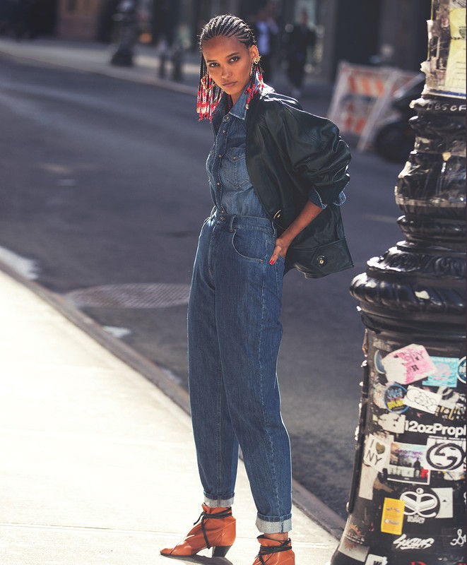A stylish Black woman in a denim jumpsuit and black jacket poses on a city street with a vintage-style lamppost.