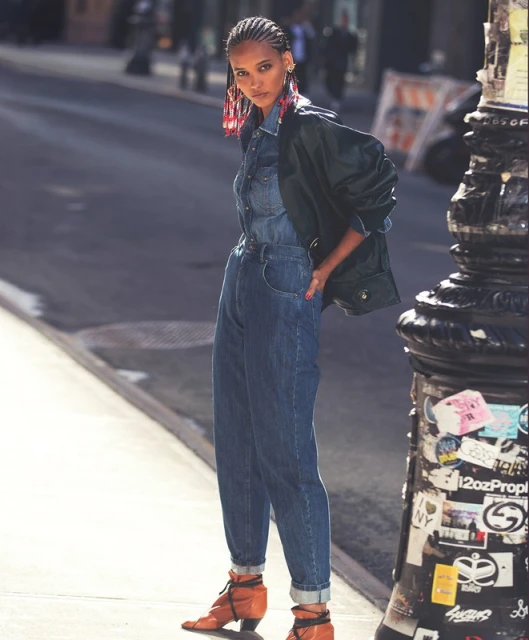 A stylish Black woman in a denim jumpsuit and black jacket poses on a city street with a vintage-style lamppost.