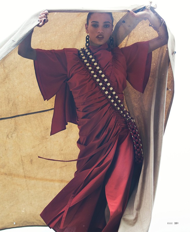 Woman in a maroon dress holds up a beige tent flap, posing in a bright, outdoor setting with fashion-forward style and editorial feel.