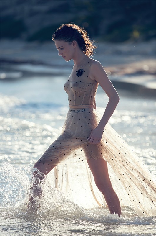 Woman wearing sheer polka dot dress wading in ocean water with splashing waves, photographed by David Bellemere.