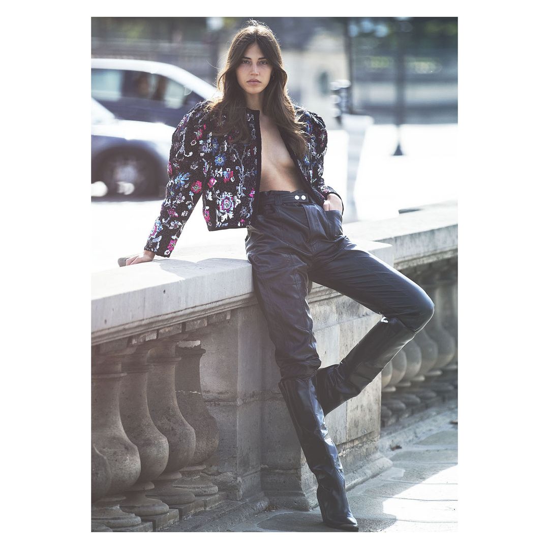 Fashion shot of a slender brunette posing in black leather pants and high-heeled boots and patterned cropped jacket in an urban setting.