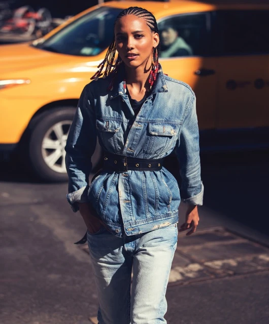 Woman in double denim and a black belt walks on a city street with a yellow taxi in the background on a sunny day.