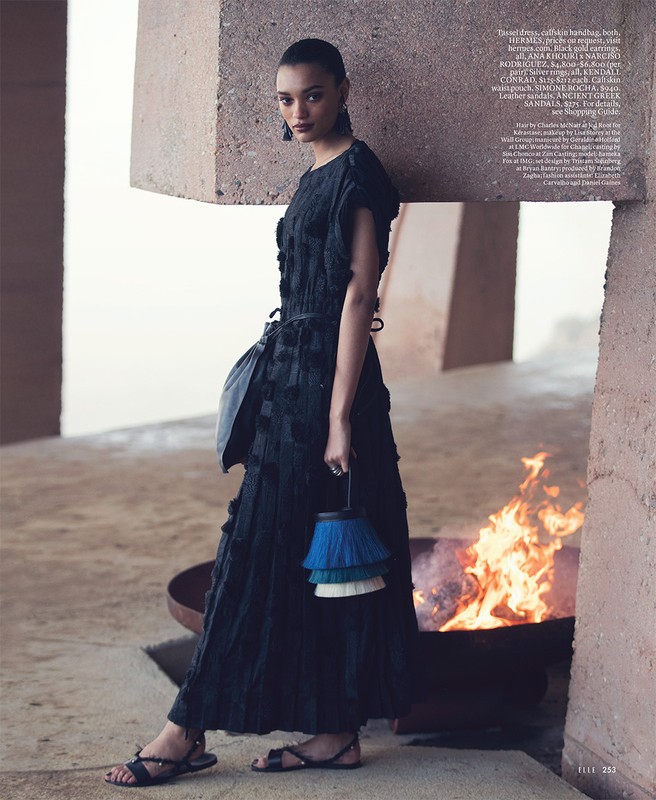 Fashion editorial shot of a woman in a dark dress and sandals holding a purse standing next to a fire pit outdoors.