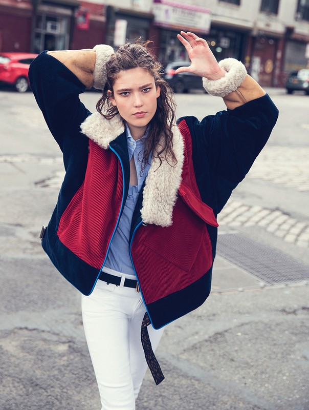 Woman in color-block jacket, white pants poses on city street. Jacket has shearling collar and cuffs. Urban fashion editorial shot.