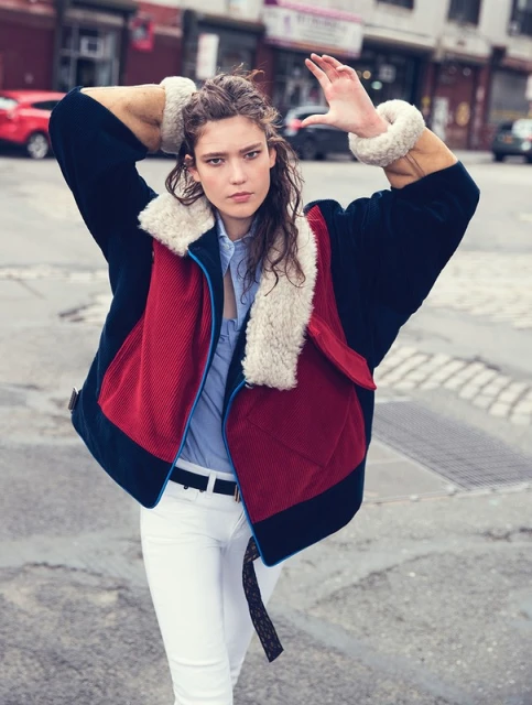Woman in color-block jacket, white pants poses on city street. Jacket has shearling collar and cuffs. Urban fashion editorial shot.