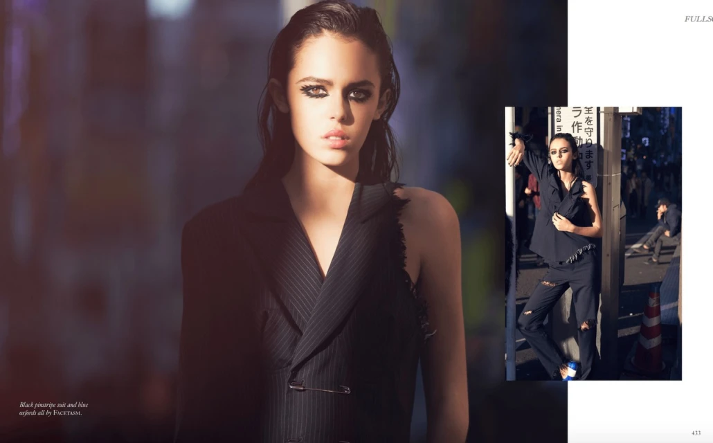Fashion editorial shot of a young woman with heavy eye makeup wearing a pinstripe jacket in an urban setting with an edgy, street-style look.