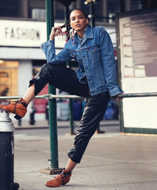 Fashionable woman in denim jacket and black pants poses on city street, showcasing trendy leather ankle boots and braided hairstyle.