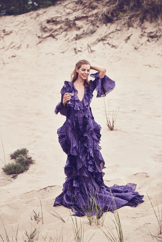 Woman in a long purple ruffled dress holding a glass while standing on sandy dunes outdoors.