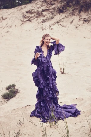Woman in a long purple ruffled dress holding a glass while standing on sandy dunes outdoors.
