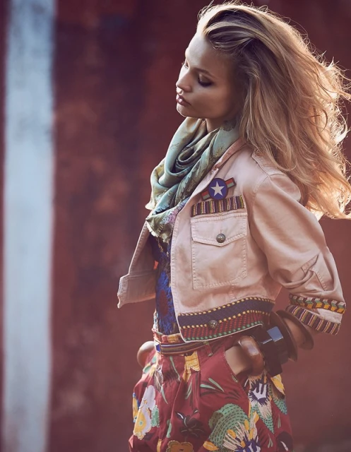 Fashion shot of a blonde woman in a stylish, light jacket and floral skirt, posing against a blurred background with soft, diffused lighting.