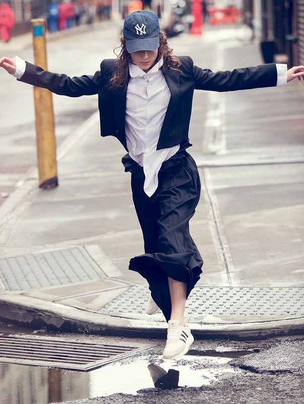 Young adult woman in a blazer, white shirt, and cap jumping over a puddle in an urban environment with arms outstretched.