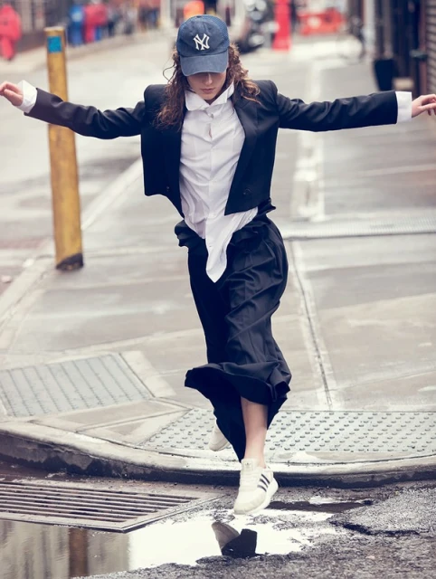 Young adult woman in a blazer, white shirt, and cap jumping over a puddle in an urban environment with arms outstretched.