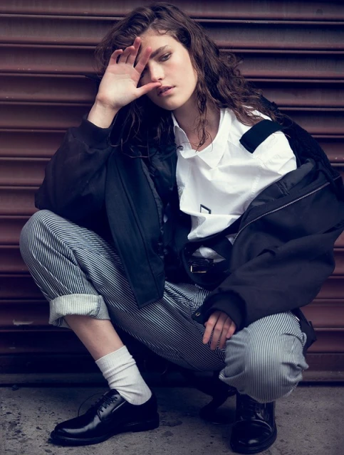 A young adult woman crouches outdoors against a metal wall wearing a white shirt, striped pants, black jacket, and black dress shoes.