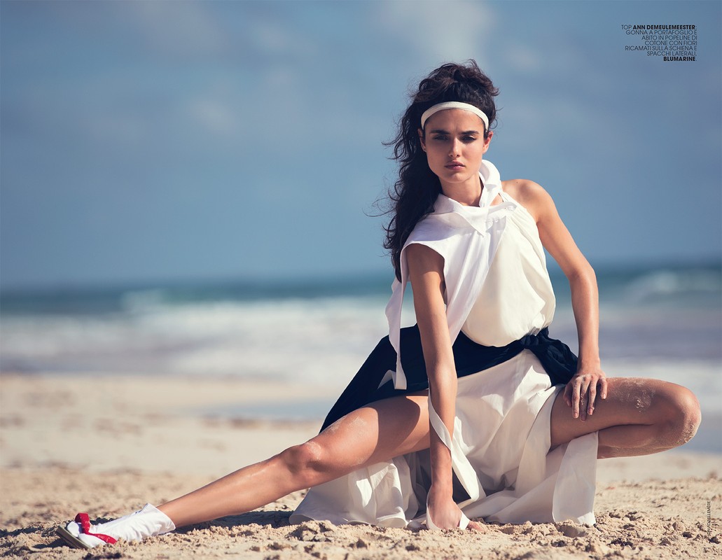 Adult woman in white dress poses in the sand on a sunny beach with ocean and sky in the background in an outdoor fashion shot.