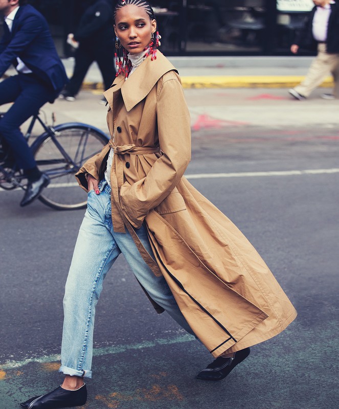 Woman in a beige trench coat and light denim jeans walking on a city street by David Bellemere
