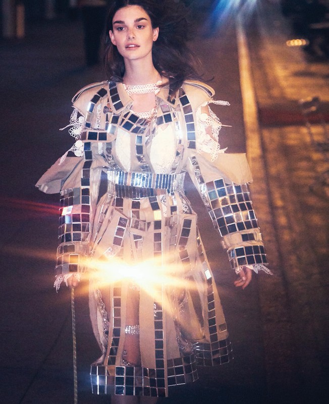 A model wearing a tan trench coat decorated with small mirrors poses against a dark background with light flares for a fashion shoot.