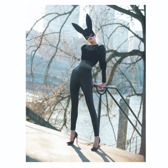 Fashionable woman in bunny mask and black outfit poses confidently outdoors against a city background, wearing heels and holding a railing.