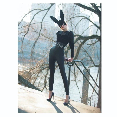 Fashionable woman in bunny mask and black outfit poses confidently outdoors against a city background, wearing heels and holding a railing.