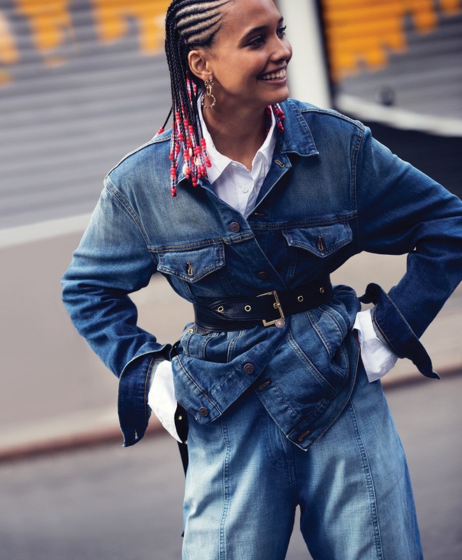 Smiling woman in a denim jacket and wide-leg jeans poses outside with braided hair featuring red beads, creating a stylish fashion moment.