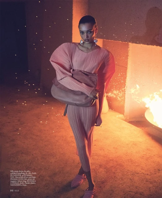 Editorial fashion shot of an adult female model in a blush-toned pleated dress, carrying a leather handbag, in front of a fire pit.