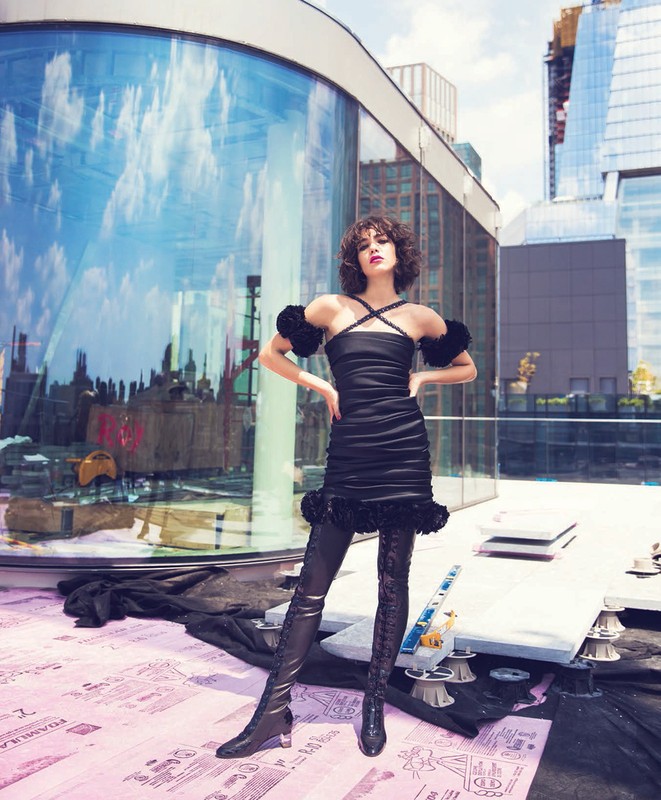 Young woman in stylish black dress and thigh-high boots poses on a rooftop against a backdrop of modern buildings and blue sky.