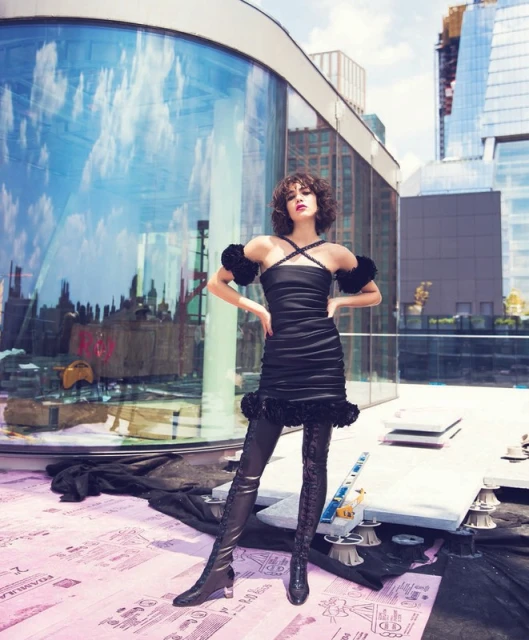 Young woman in stylish black dress and thigh-high boots poses on a rooftop against a backdrop of modern buildings and blue sky.