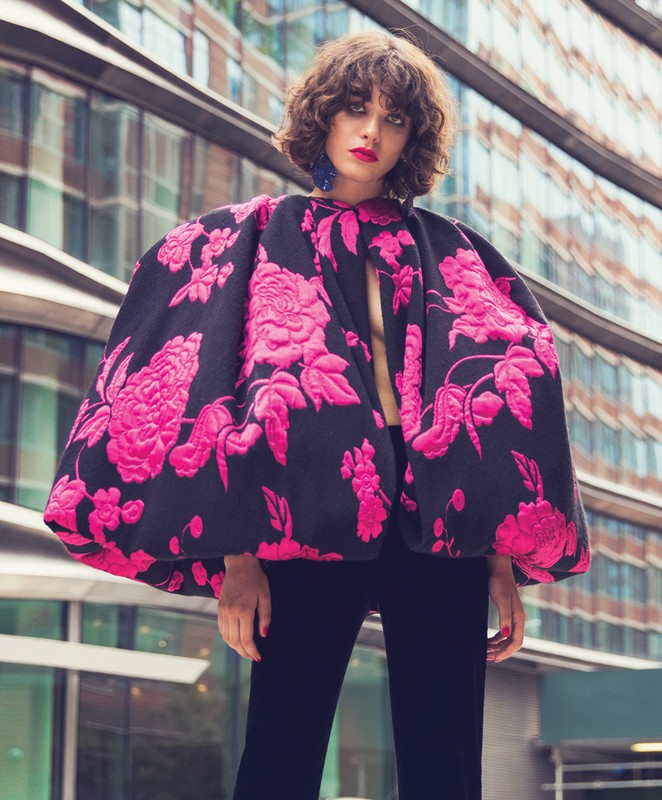 Fashionable adult woman poses outdoors in a black and bright pink floral print jacket and black slacks with a modern building backdrop.