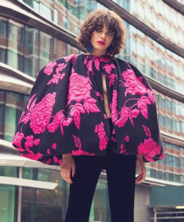 Model with curly brown hair wearing a voluminous black and pink floral cape against a modern office building exterior