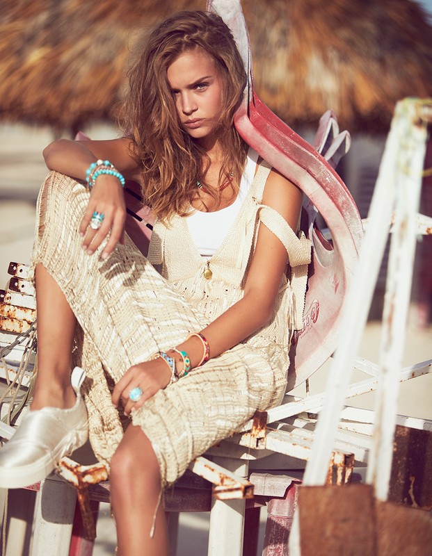 A fashionable adult woman sits and poses outdoors, wearing a white top, knit dress, jewelry, and sneakers, against a blurry background.