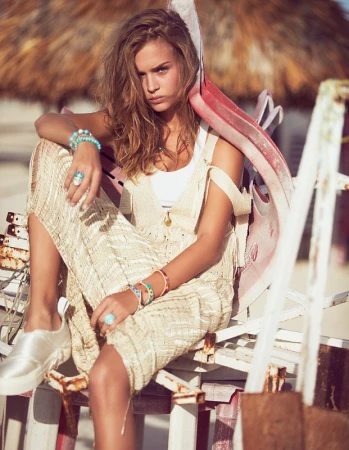A fashionable adult woman sits and poses outdoors, wearing a white top, knit dress, jewelry, and sneakers, against a blurry background.
