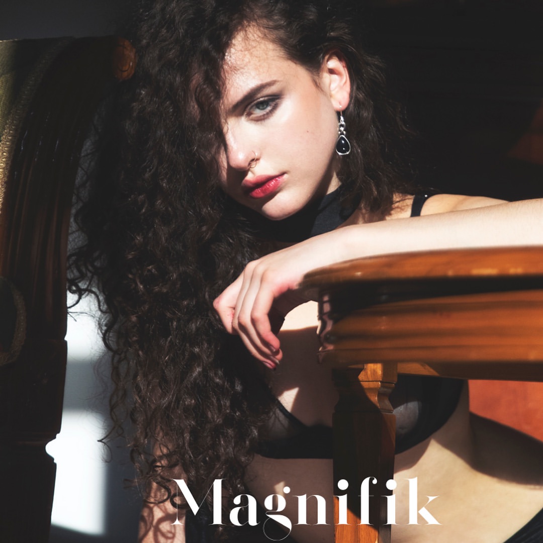 Brunette woman with long curly hair poses indoors wearing a black bra and top in natural lighting, next to wooden table.