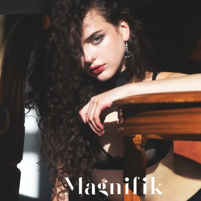 Brunette woman with long curly hair poses indoors wearing a black bra and top in natural lighting, next to wooden table.