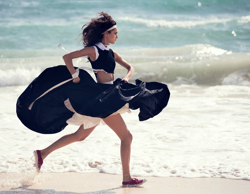 A woman in a black and white dress runs along a sandy beach next to the ocean on a bright, sunny day with a playful expression.