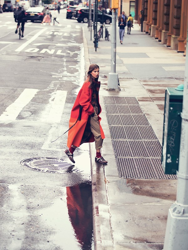 A woman in a red coat crosses a wet city street with puddles reflecting buildings. Pedestrians and cars are in the background.