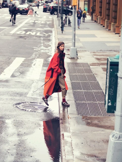 A woman in a red coat crosses a wet city street with puddles reflecting buildings. Pedestrians and cars are in the background.