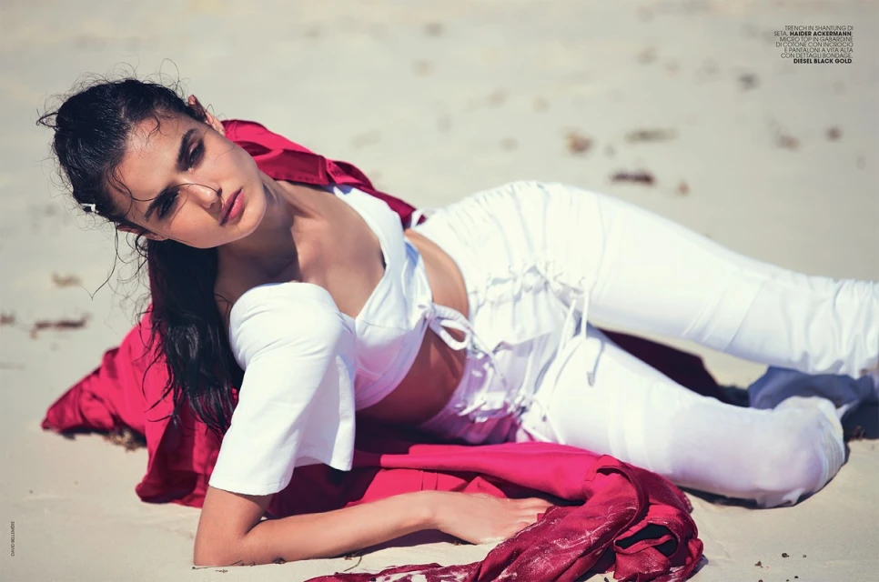 A woman in a white outfit and red shirt lays on a sandy beach on a sunny day, with her head slightly tilted and looking off to the side.