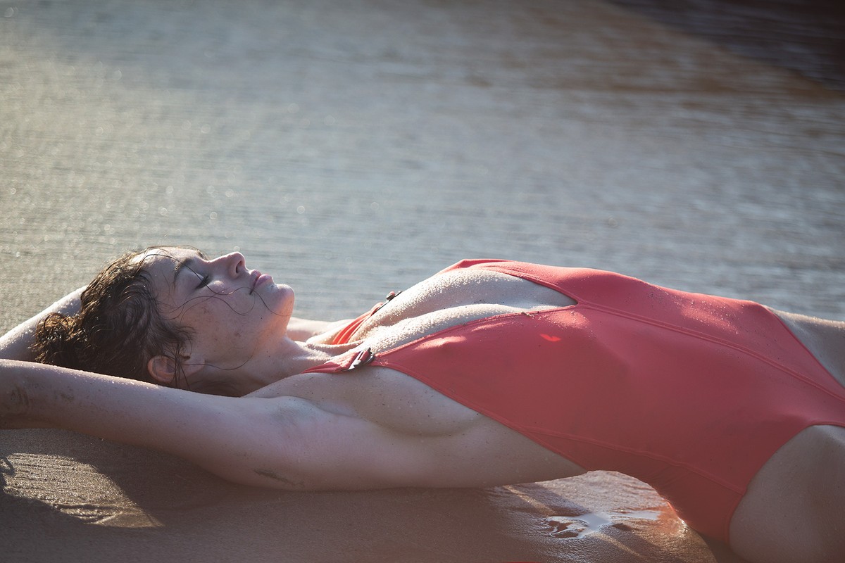 Woman in swimsuit lying on beach with wet sand, sunlit, looking relaxed and serene in outdoor daylight.