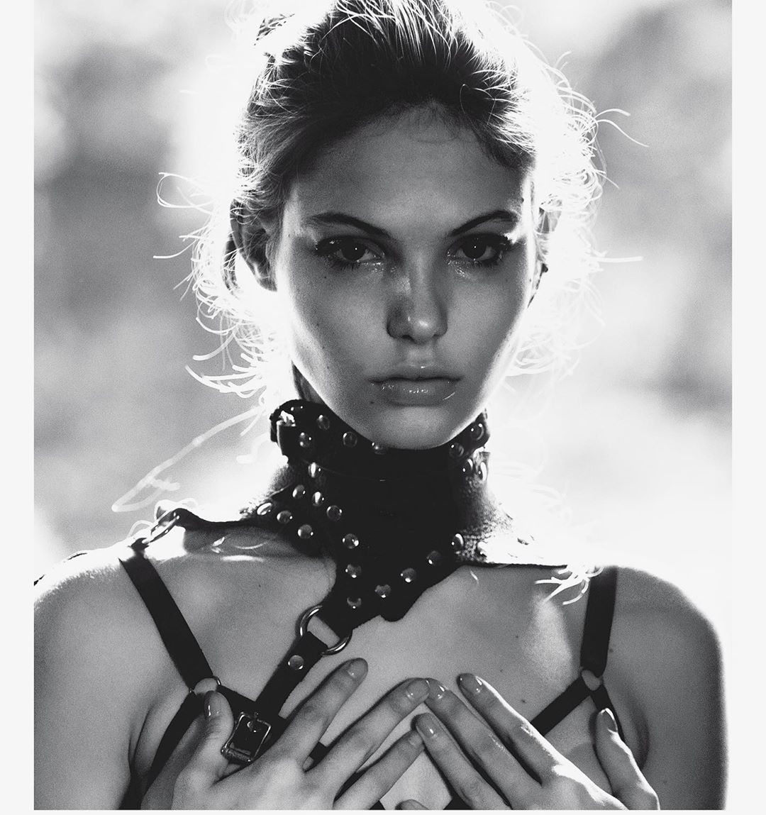 Monochrome portrait of a young woman with fair skin, hands raised to her chest, wearing a studded leather harness and choker.