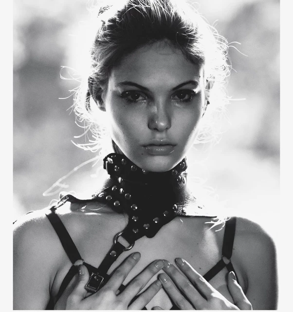 Monochrome portrait of a young woman with fair skin, hands raised to her chest, wearing a studded leather harness and choker.